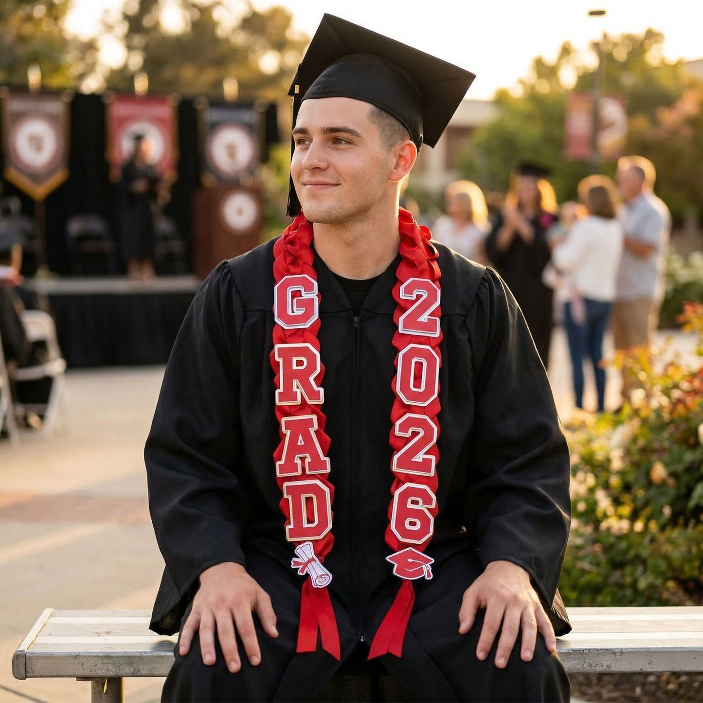 A smiling male graduate wearing a black cap and gown with a custom red and silver braided graduation lei. The lei features "GRAD" and "2026" foil cardstock accents. The graduate is seated outdoors at a commencement ceremony during golden hour.