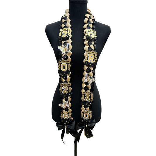 Handcrafted 2026 graduation ribbon lei featuring a black and gold military braid design, accented with 3D white and gold butterflies, gold "GRAD" and "2026" lettering, and black satin bows on a mannequin.