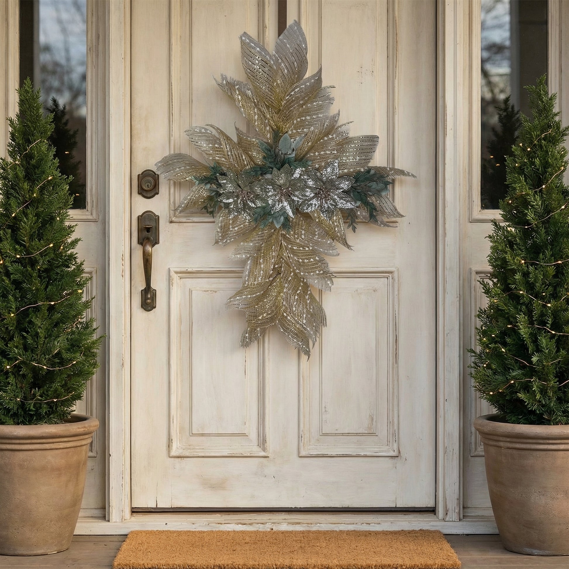 A lifestyle photograph of a glittered burlap cross wreath hanging on a distressed off-white wooden front door. The wreath features silver-glittered poinsettias and gold-striped mesh. The entryway is flanked by two tall evergreen topiary trees in rustic clay pots wrapped in warm micro-fairy lights, with a simple tan coir doormat at the base.