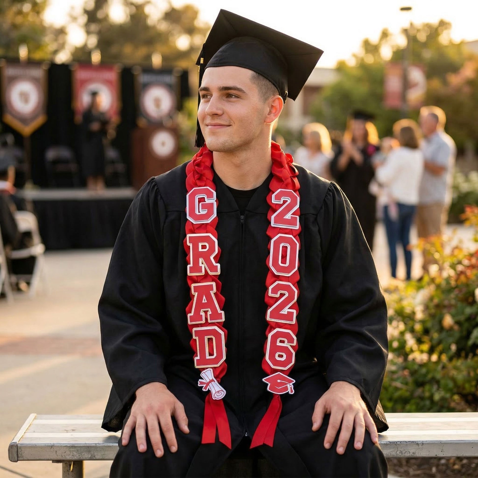 A smiling male graduate wearing a black cap and gown with a custom red and silver braided graduation lei. The lei features "GRAD" and "2026" foil cardstock accents. The graduate is seated outdoors at a commencement ceremony during golden hour.