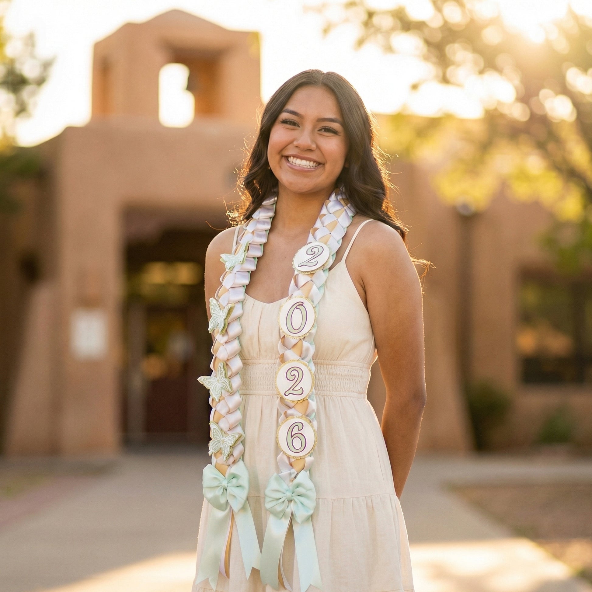 professional outdoor senior portrait of a smiling graduate wearing a cream-colored sundress and a handcrafted 2026 graduation lei featuring a mint green, gold, and white military braid with butterfly accents.