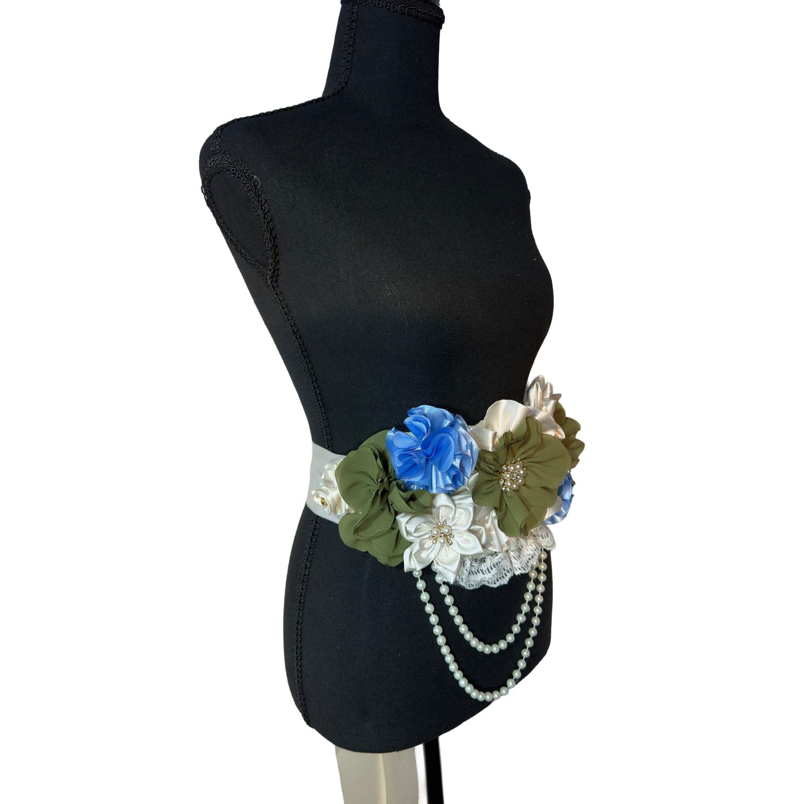Angled view of a green and light blue maternity flower belt with rhinestone centers and delicate white lace detailing.