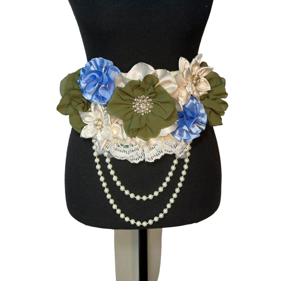 Front view of a handmade baby shower sash featuring green chiffon flowers, blue satin rosettes, and draped faux pearls.