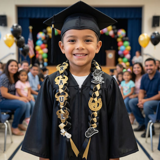 A 5-year-old boy smiling in a black graduation gown and cap, wearing a custom handcrafted black and gold western-themed satin ribbon graduation lei for kindergarten.