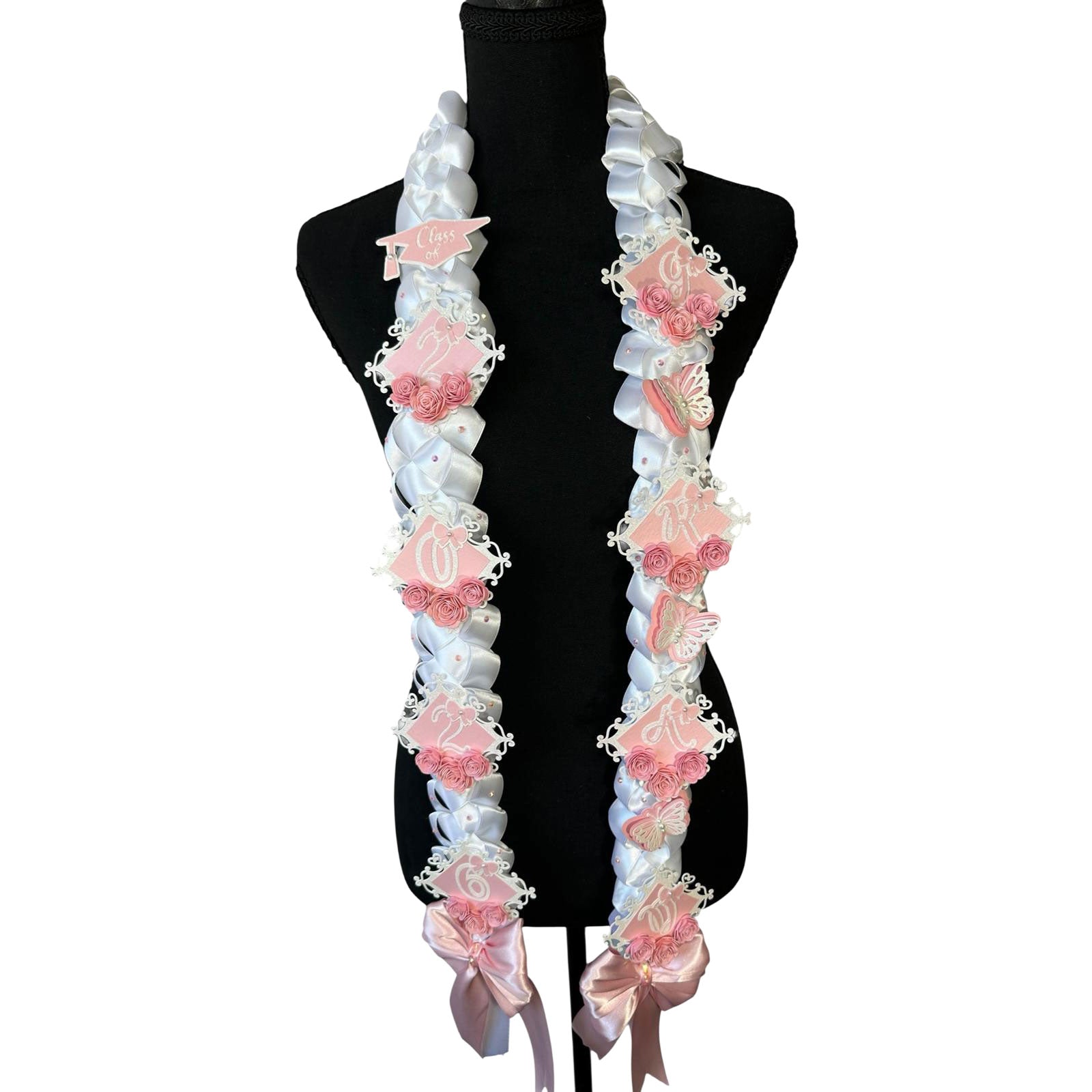 Front view of a 2026 graduation lei featuring a white single military braid design with soft pink butterfly accents, pink paper roses, and personalized pink glitter lettering on a black display mannequin.