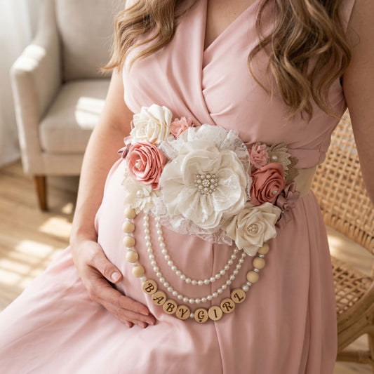 A handmade Mommy-to-Be sash featuring dusty rose, blush, and ivory fabric flowers with a large lace peony center. The sash includes a personalized beaded fringe with white pearls, wooden beads, and a custom name charm spelling "Baby Girl" against a soft white background.
