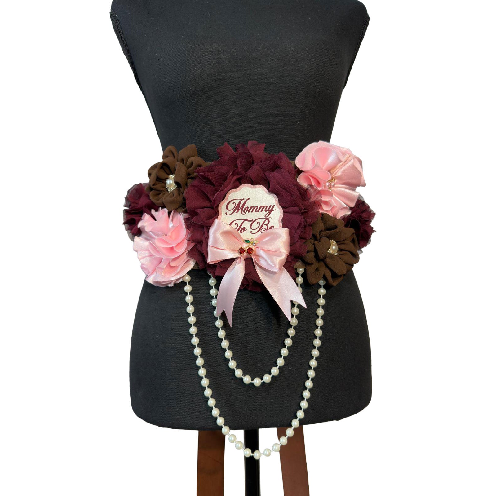Front view of a maternity belly sash on a black mannequin featuring layered chiffon and satin flowers in burgundy, pink, and chocolate brown with draped pearl strands.