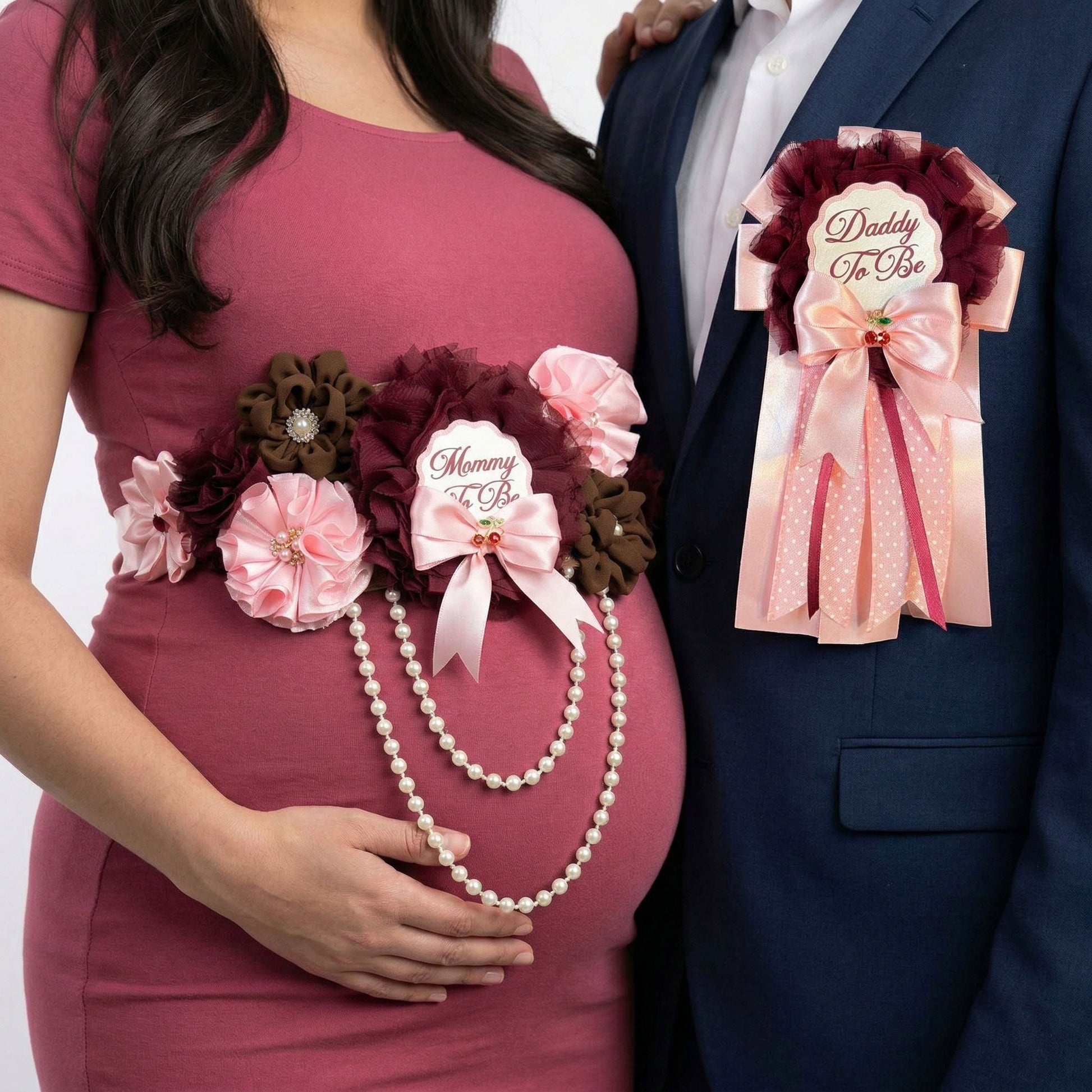 Lifestyle shot of a pregnant woman wearing a burgundy and pink floral maternity sash and a man in a navy suit wearing a matching Daddy-to-Be pin.
