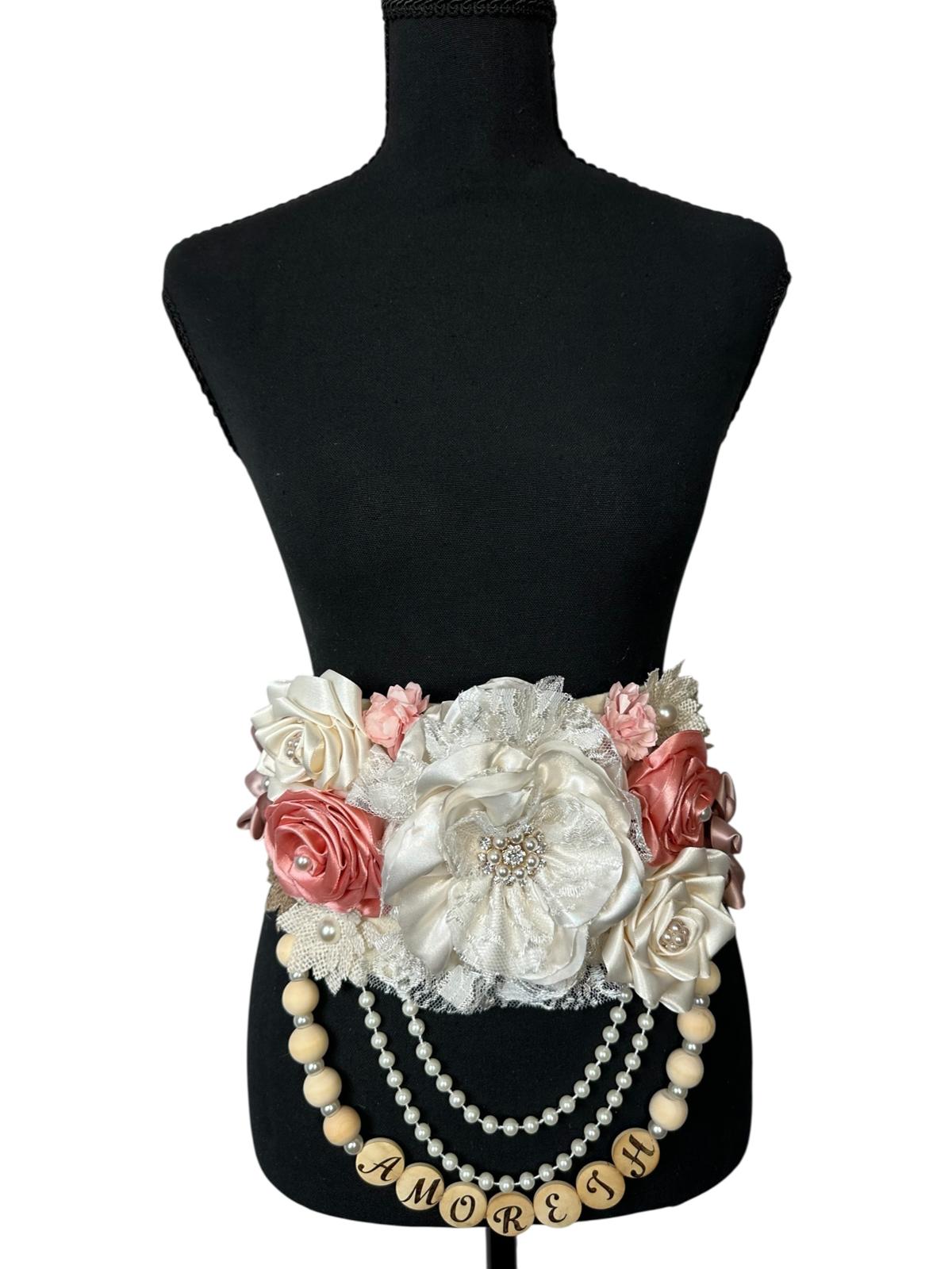 Full view of a custom "Mommy-to-Be" maternity sash featuring a cluster of blush, dusty rose, and ivory fabric flowers with a personalized beaded name charm spelling "Amoreth" on a white background.