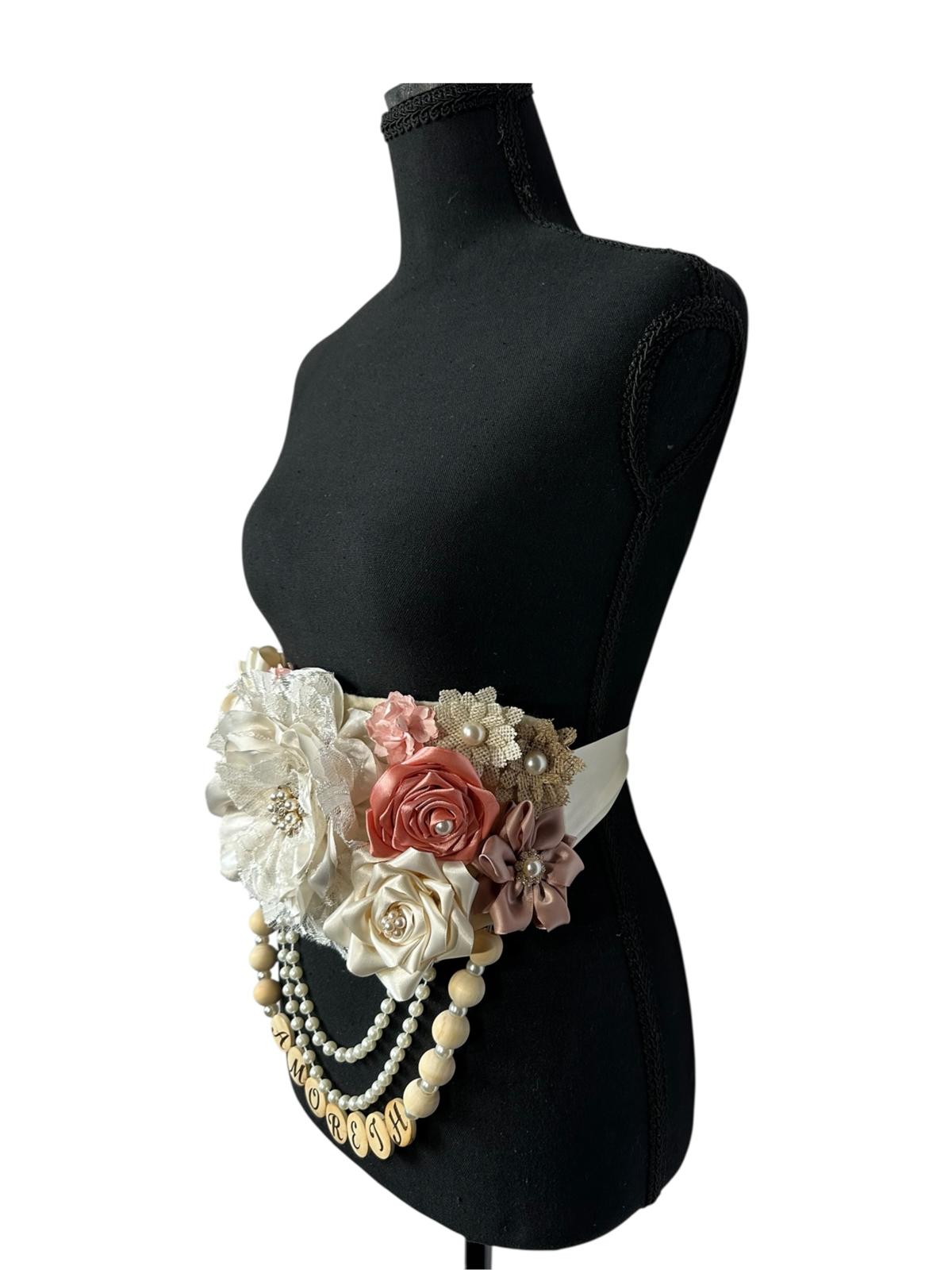 Close-up detail of the floral arrangement on a personalized maternity sash, showing a large ivory lace peony with a rhinestone center, surrounded by satin rosettes, burlap accents, and delicate pearl sprays.
