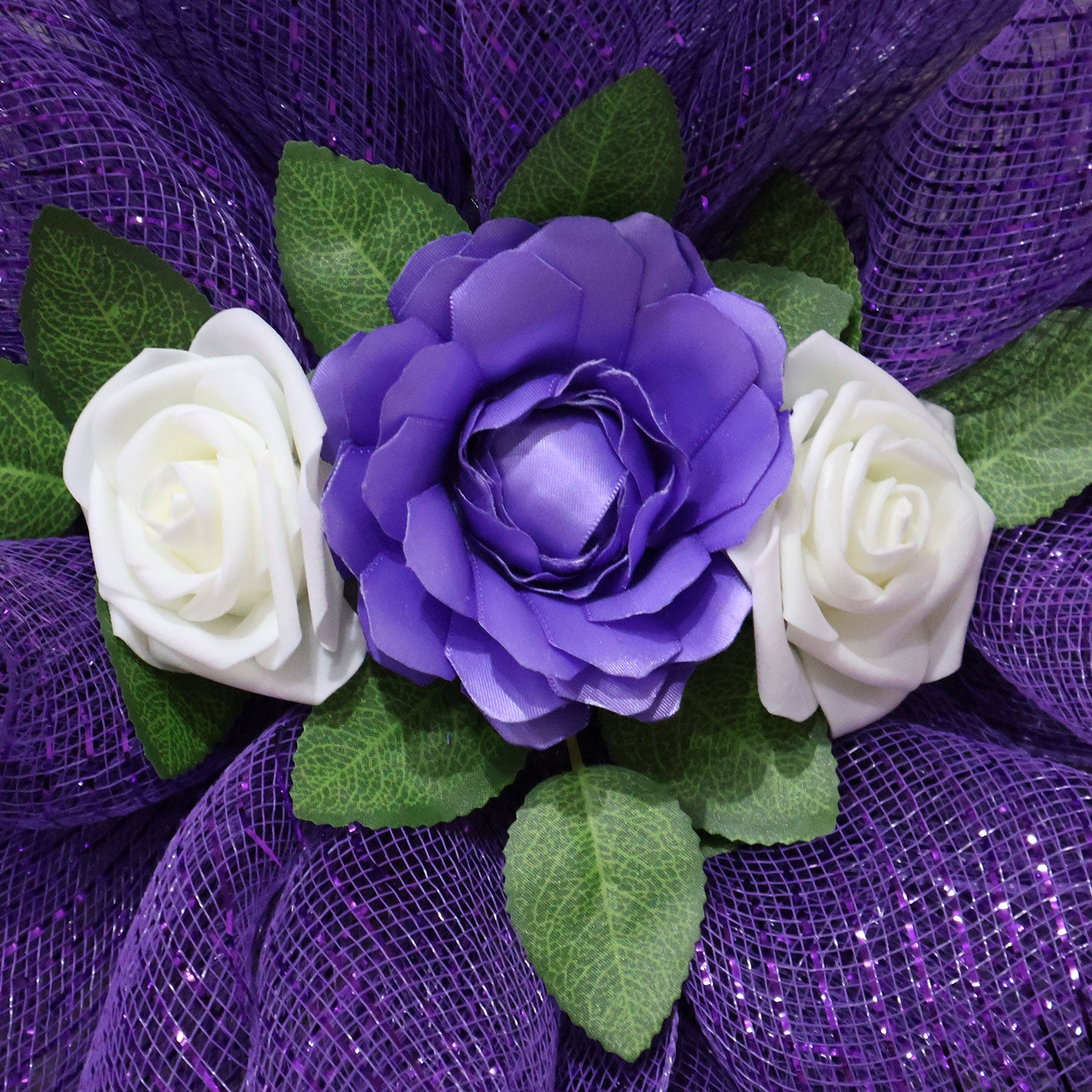 Close-up detail of a cross wreath showing a handmade lavender double-satin ribbon rose, two off-white artificial roses, and realistic green leaves nestled in purple deco mesh.