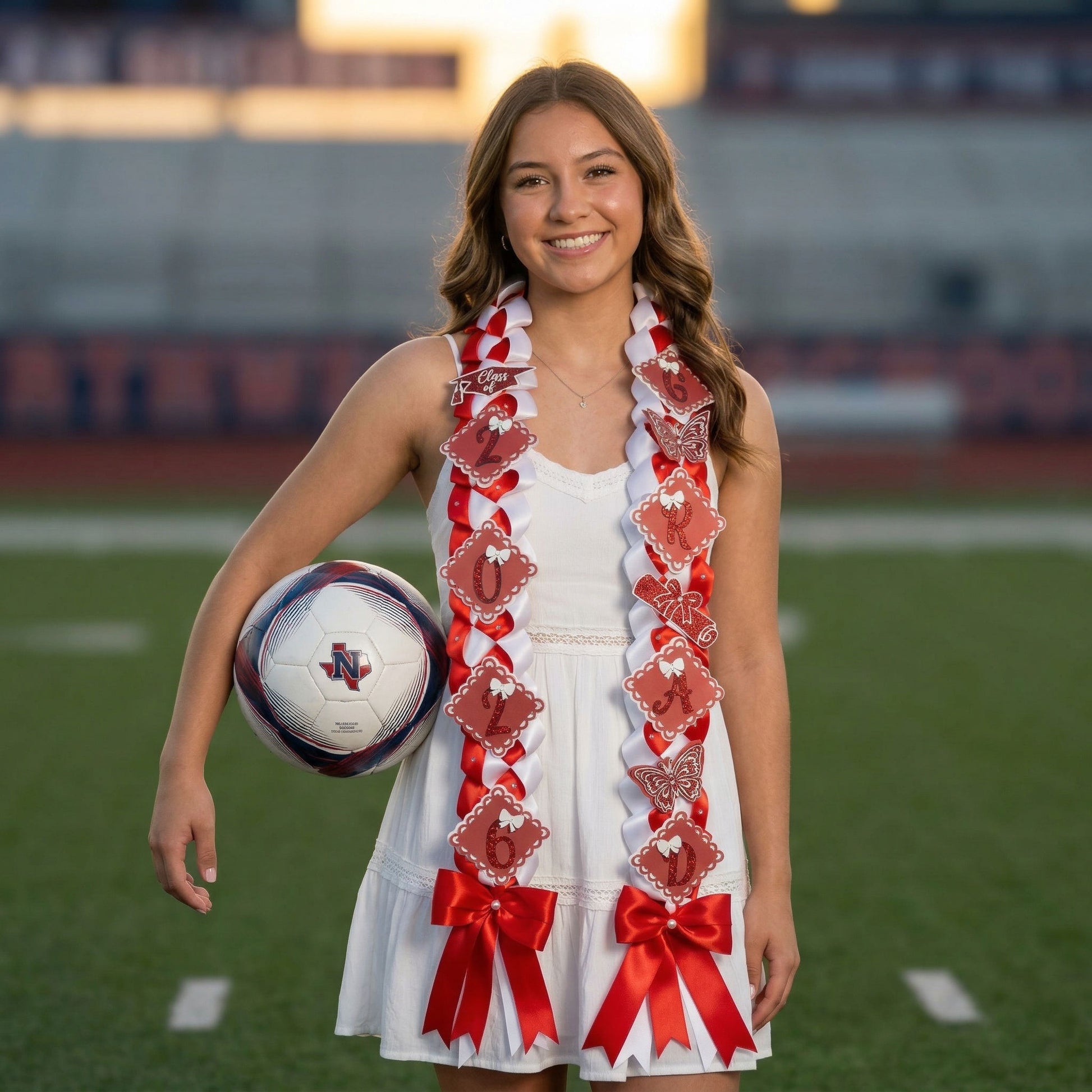 A female high school graduate in a white dress and graduation cap stands on a soccer field holding a soccer ball, wearing a red and white military braid graduation lei with "GRAD" and "2026" glitter accents. The background is a soft-focus athletic field at sunset, highlighting the Northwest High School "Texan" team colors.