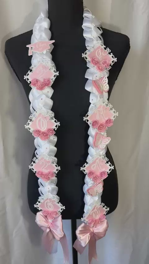 A close-up video showcasing a handcrafted 2026 White & Pink "Butterfly" Graduation Lei. The video pans along the white satin military braid, highlighting the shimmering pink glitter GRAD lettering, delicate pink paper roses with rhinestone centers, and layered pink and white butterflies that flutter with movement.