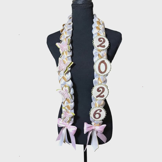 A 360-degree view of a 2026 graduation lei featuring white and gold braided ribbons. One side is adorned with delicate pink and white butterflies, while the other features scalloped circles displaying the graduation year '2026' in gold. The lei is finished with elegant pink and white bows at the bottom.